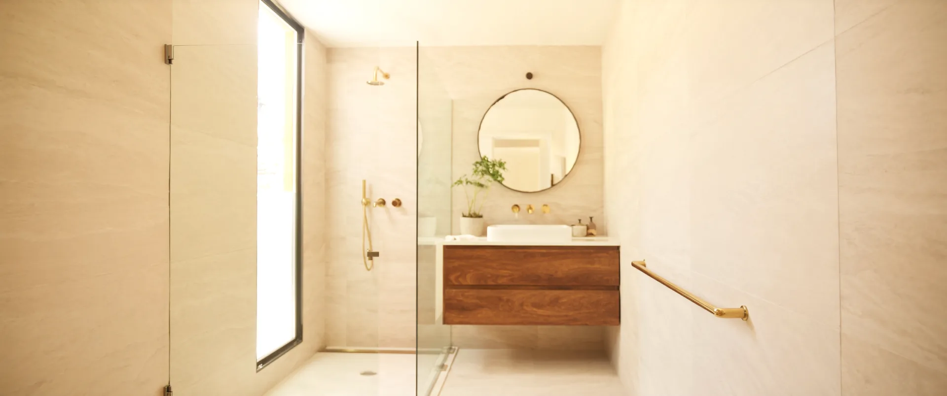 A narrow compact bathroom in warm cream stone tile, with a frameless glass walk-in shower lit by a tall window, a walnut wall-hung vanity and a round mirror