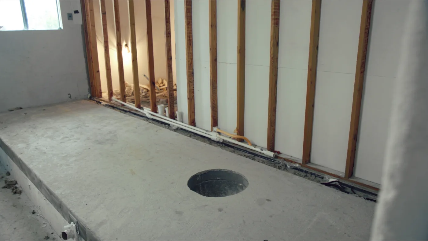 Bare concrete slab in a bathroom mid-renovation, with the toilet drain opening cut into the slab and plumbing rough-in in the open wall framing