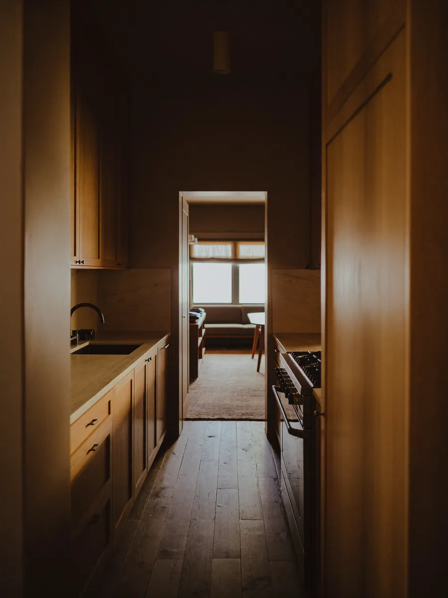 A compact galley kitchen in warm wood and pale stone, two facing runs of cabinets with a narrow walkway between them, lit by a window at the far end