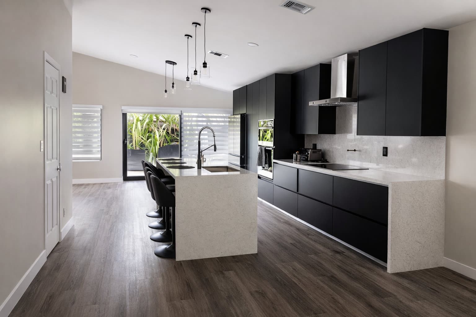 Single-family kitchen remodel with matte black flat-front cabinetry, light stone waterfall island, stainless range hood, and casement door to planted exterior, Miami-Dade County