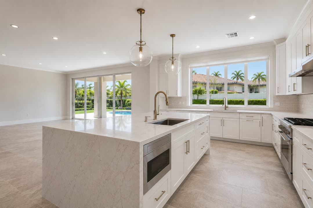 Single-family kitchen remodel with white shaker cabinetry, marbled stone waterfall island, brushed brass hardware and glass globe pendants, bright open coastal atmosphere, Miami-Dade County