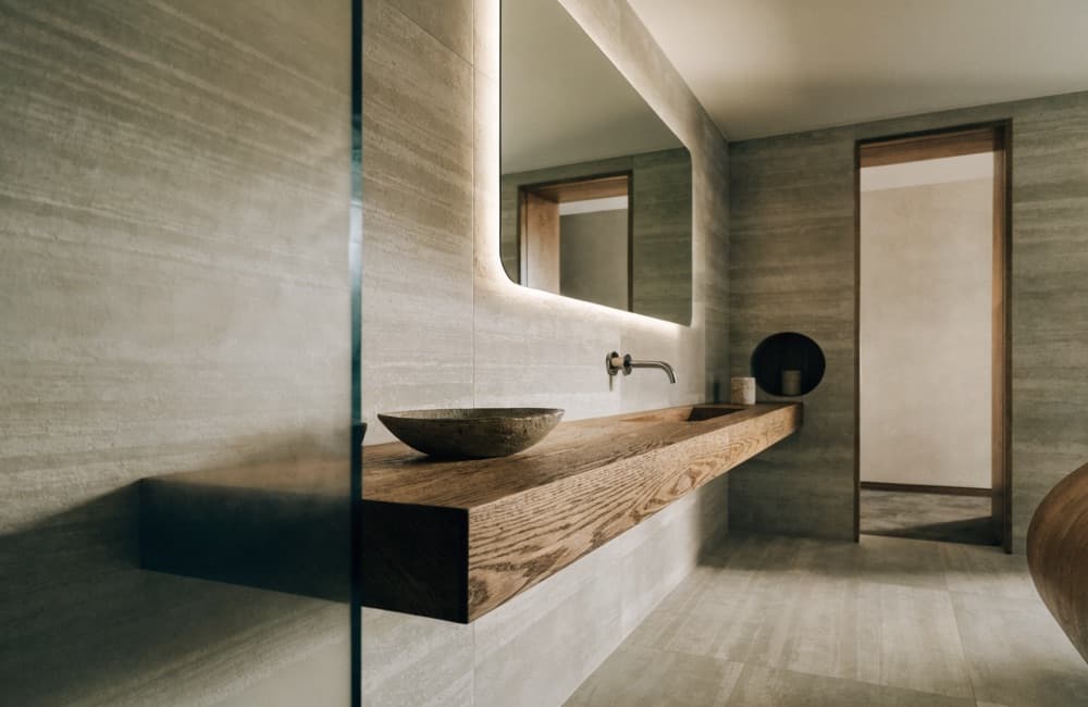 Finished bathroom with large-format stone-look tile, a floating wood vanity shelf, a vessel basin and a backlit mirror.