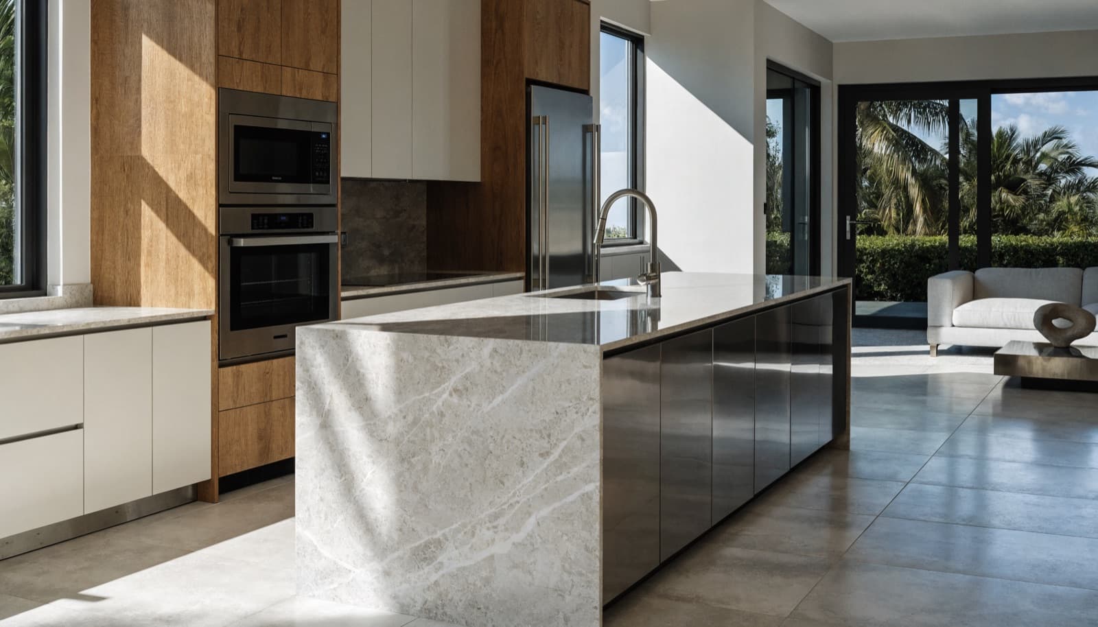Kitchen with a stone waterfall island, wood and white cabinetry with built-in ovens, and a living area opening onto palm trees.